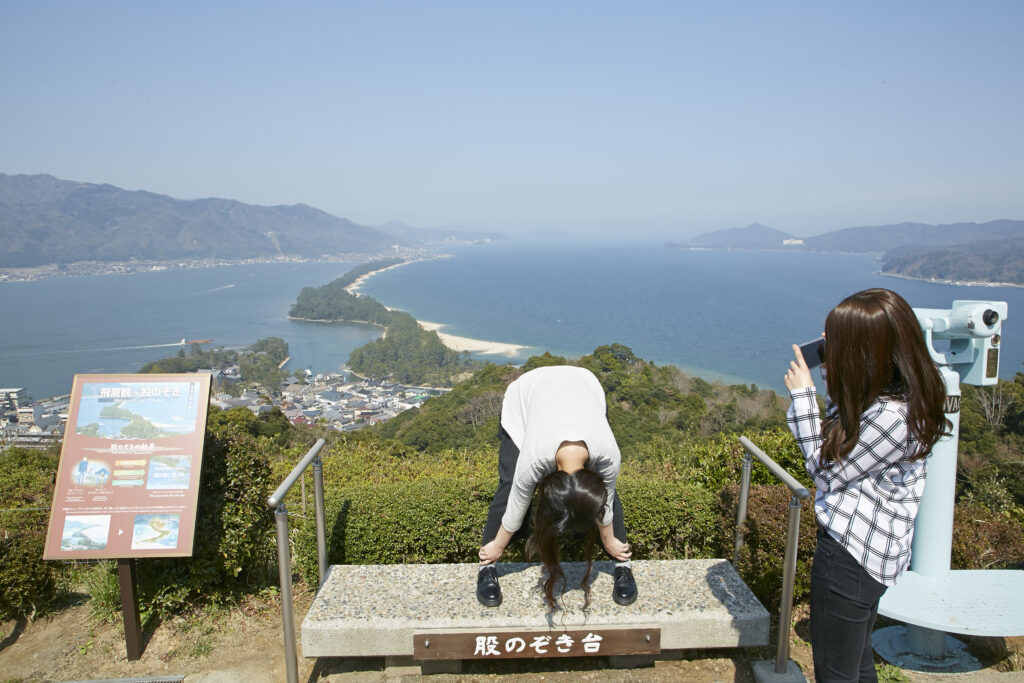 Person taking a photo of someone looking through their legs with Amanohashidate in the background