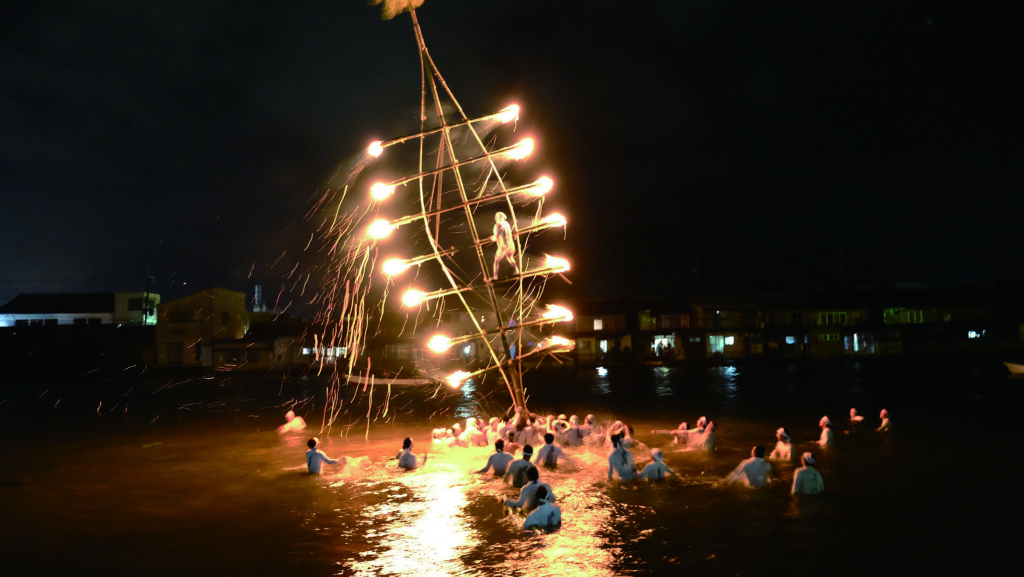 A group of people swimming in water with giant fish-shaped bamboo mast set alight