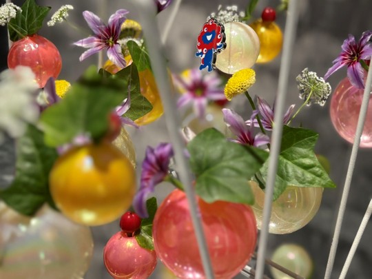 Decorative clear and colorful orbs with flowers and greenery hang, with a mini Expo 2025 mascot visible among them.