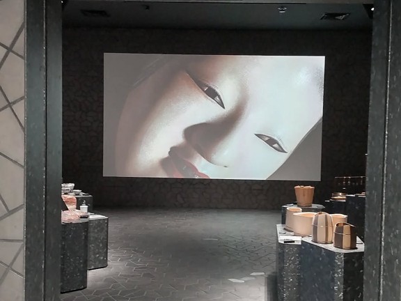 A projected image of a woman's face dominates a dark room with display pedestals featuring various ceramic and wooden items.