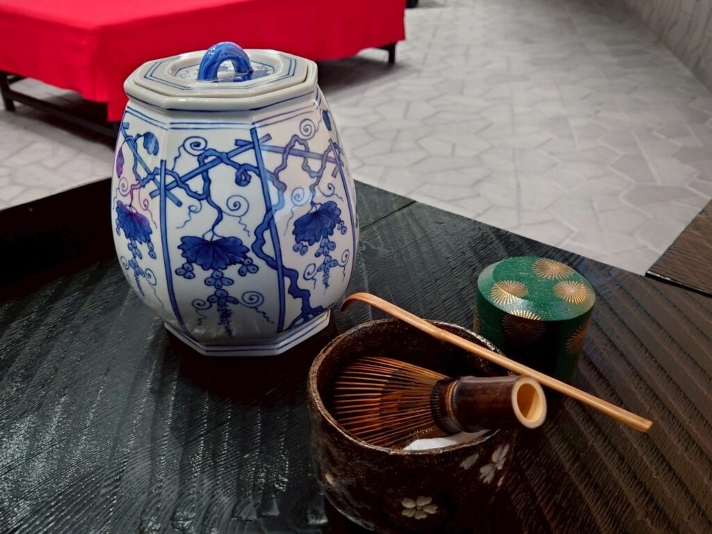 A traditional Japanese tea ceremony set, including a blue and white ceramic jar, a matcha bowl with whisk, and a small green container.