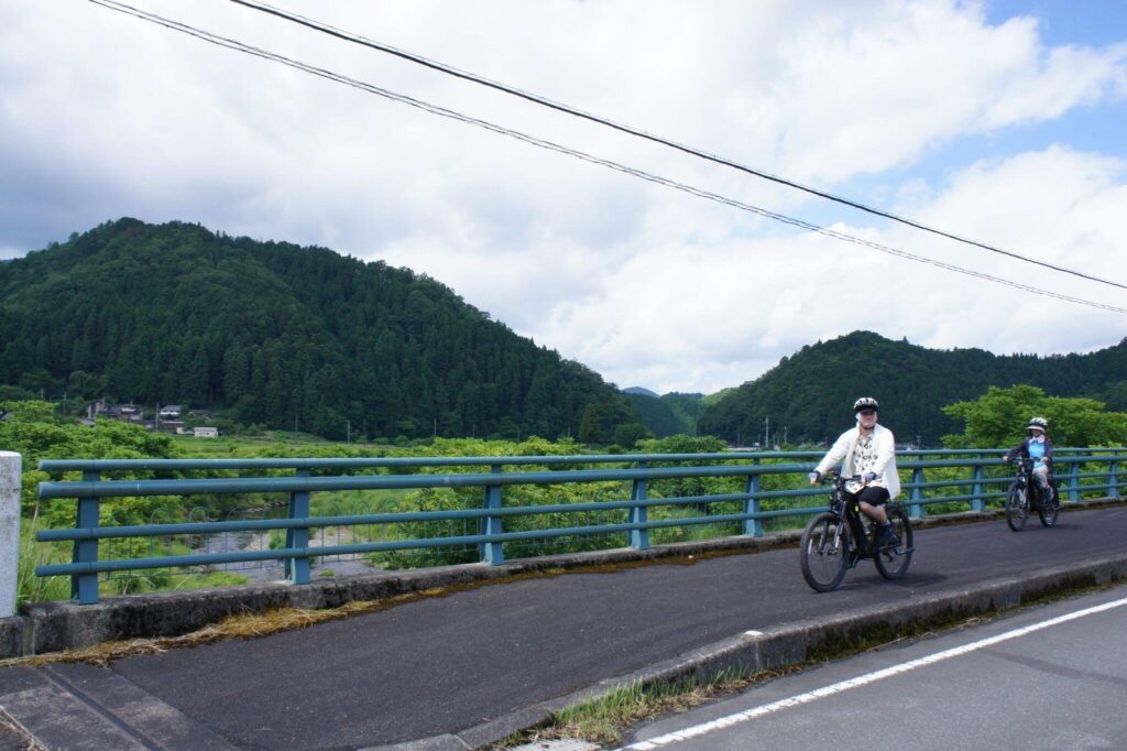 Two cyclists ride their e-bikes across a bridge, with a backdrop of Ayabe's rolling green hills and fields.