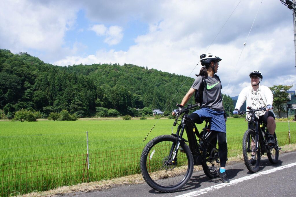 Two cyclists pause on their e-bikes to take in the view of a vast green rice paddy in the Ayabe countryside.