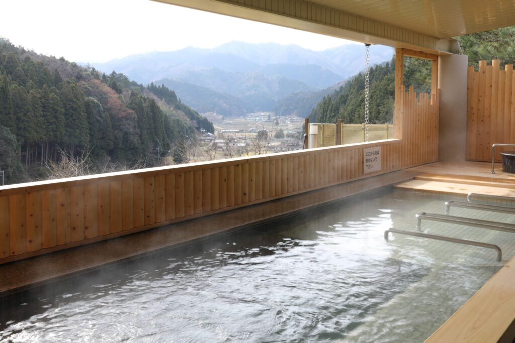 An open-air onsen in Ayabe, with steaming hot spring water and a scenic view of the mountains and valley.