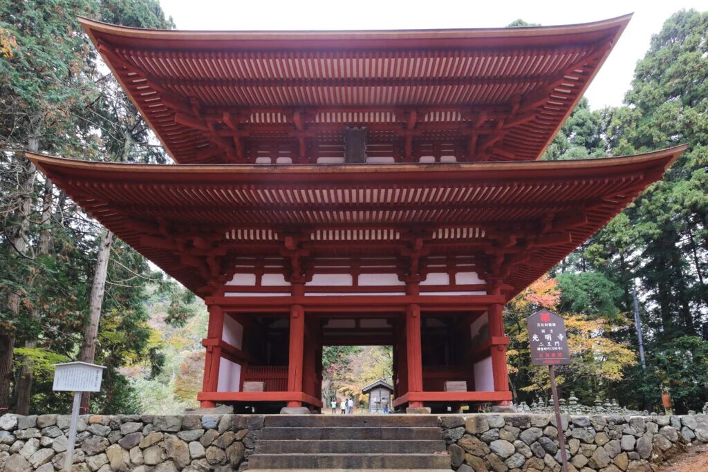 The large, two-story red Nio-mon gate of Komyoji Temple in Ayabe, set against a backdrop of dense forest.