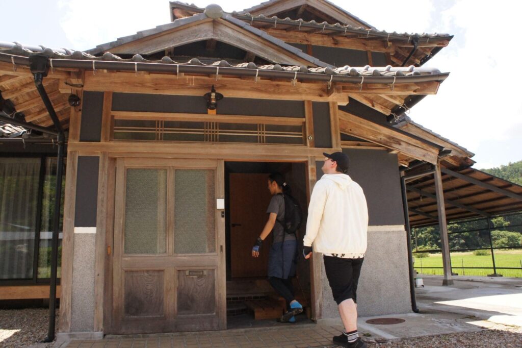 Two guests arrive and enter the Nio no Yado Biden, a traditional Japanese house with beautiful wooden architecture.